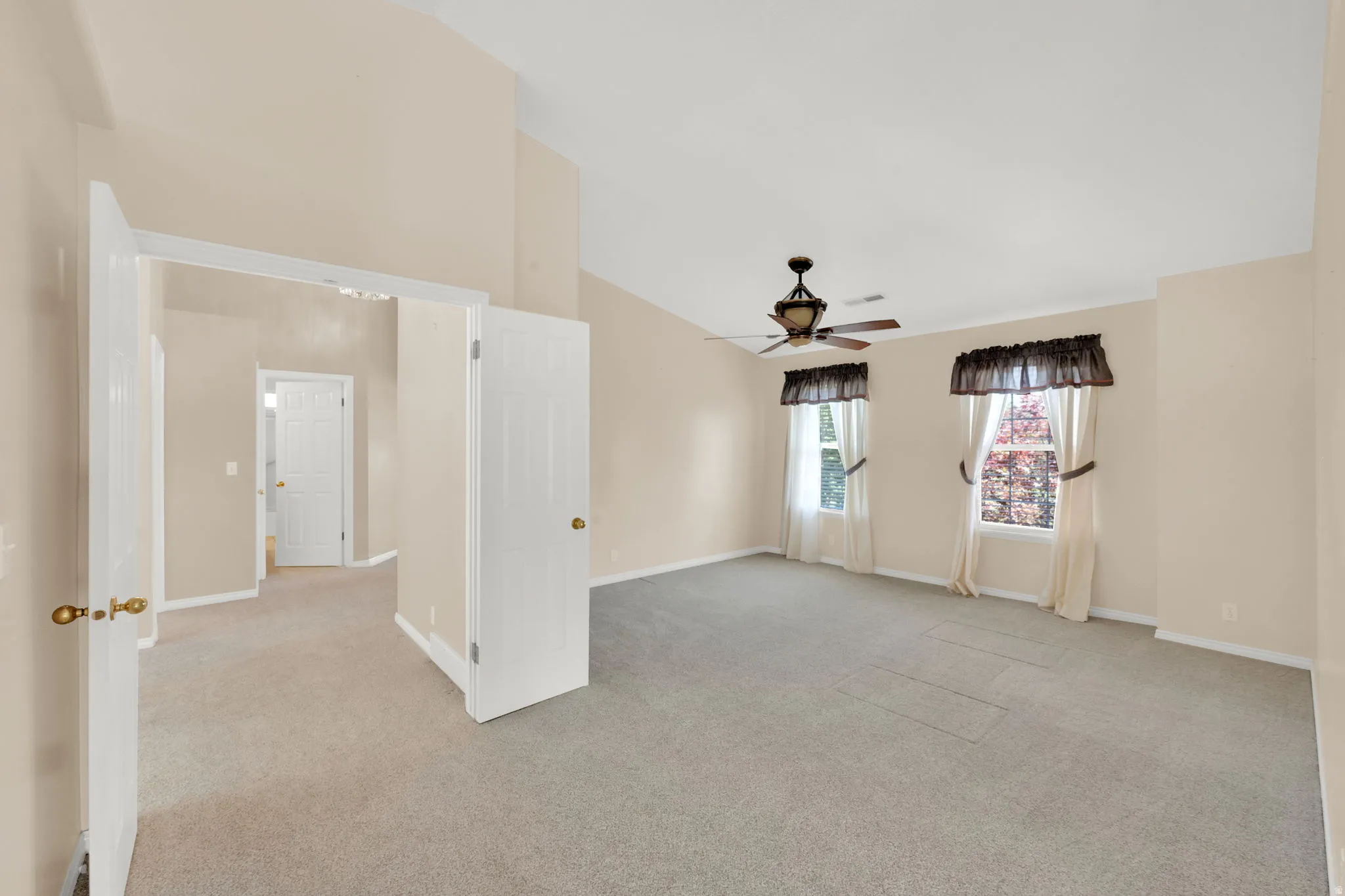 Spacious room featuring vaulted ceilings, light-colored carpeting, two large windows with valances and drapes, a ceiling fan with integrated lighting, and neutral wall paint