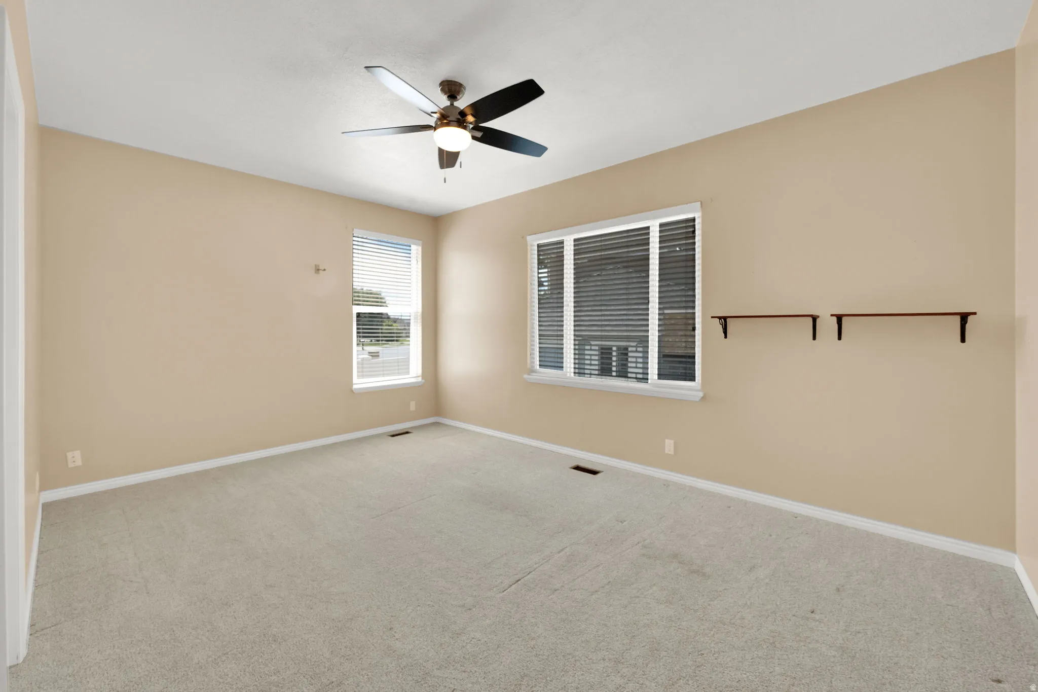Spacious carpeted room featuring a ceiling fan with integrated lighting, two windows with blinds, and built-in shelving