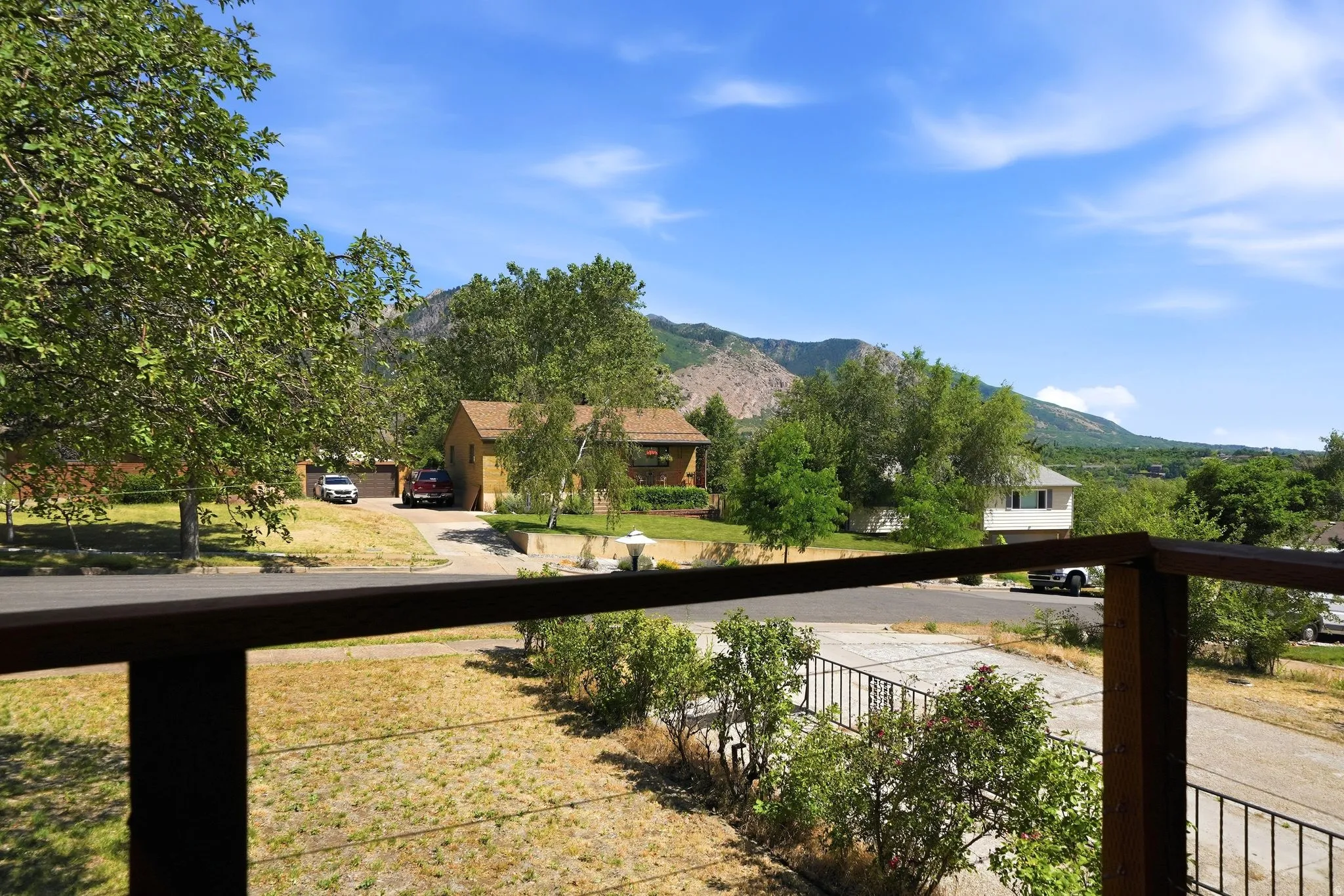 Expansive outdoor living space featuring a wood railing with cable infill, overlooking a street with mature trees and mountain scenery