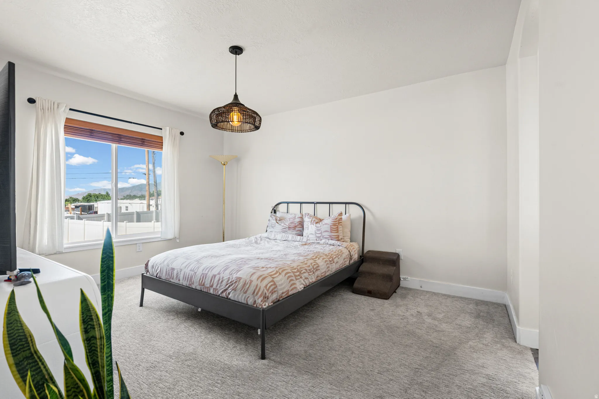 Bedroom featuring a large window with wooden blinds and white curtains, light gray carpeting, and a decorative pendant light fixture