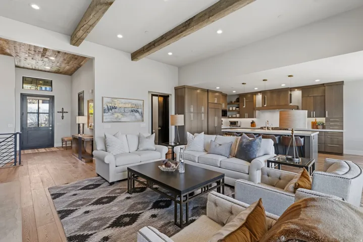 Open-concept living area featuring wood-finish flooring, exposed wood ceiling beams, recessed lighting, a modern kitchen with a large island, and a prominent entry door