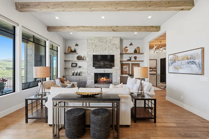 Spacious living area featuring exposed wood beams, a stone-clad fireplace with built-in shelving, large windows, recessed lighting, and wood-finish flooring