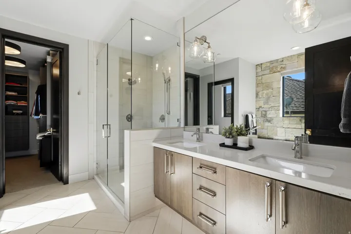 Spacious bathroom featuring a frameless glass shower enclosure, a dual vanity with undermount sinks, wood-finish cabinetry, and neutral-toned stone tile flooring