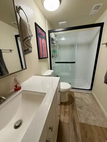 Bathroom featuring a white vanity with an integrated sink and light-colored countertop, wood-finish flooring, a toilet, and a stand-up shower with a clear glass enclosure and black trim