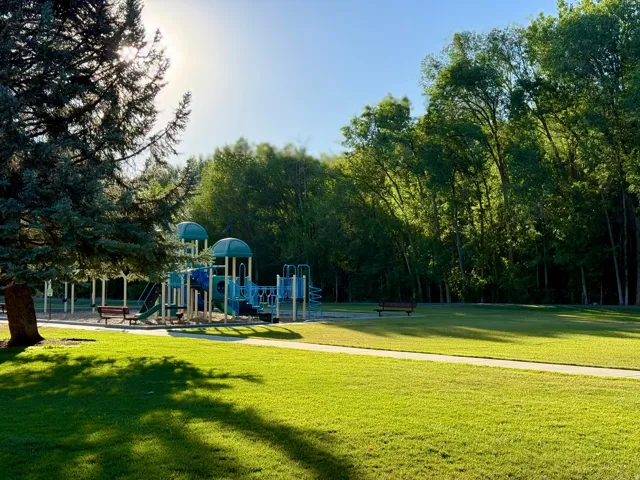 Expansive park area featuring a multi-level playground structure, mature trees, a paved walking path, and open grassy areas