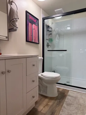 Bathroom featuring a white vanity with shaker-style cabinetry, wood-finish flooring, a contemporary toilet, and a glass-enclosed shower with white tiled walls and a sliding door