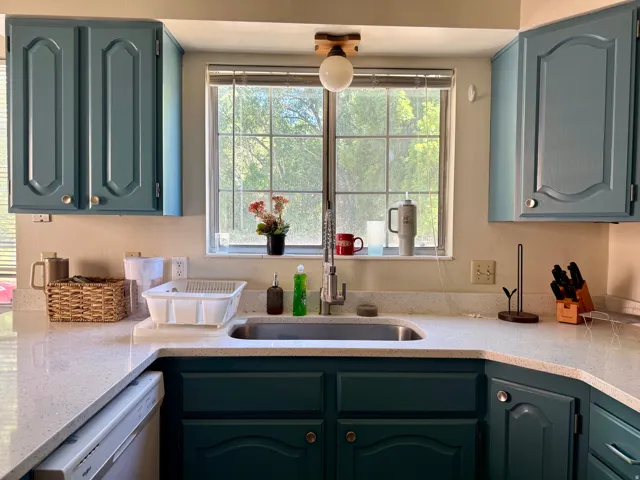 Kitchen featuring L-shaped countertops, an undermount sink with a gooseneck faucet, light-colored speckled countertops, a double-paned window, and painted cabinetry