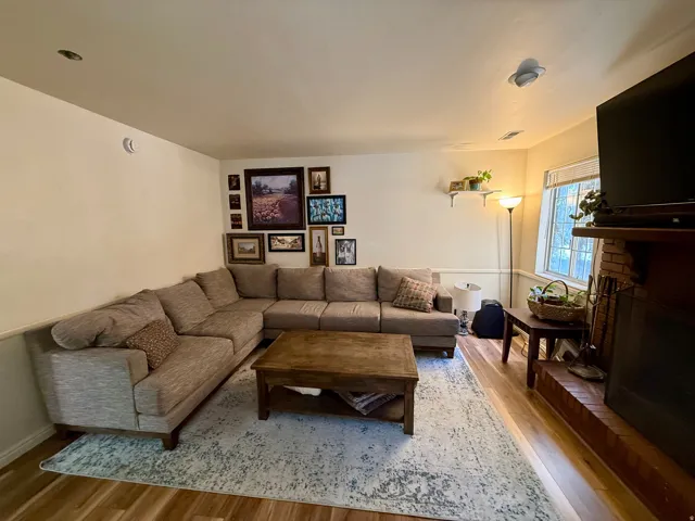 Living area featuring wood-finish flooring, a brick fireplace with a wood mantel, and a window with blinds