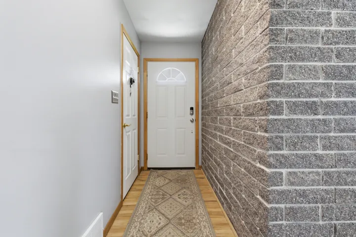 Entryway featuring a white paneled front door with an arched window insert and digital lock, complemented by light wood-finish flooring and a textured gray brick wall