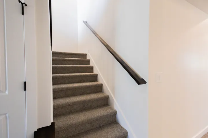 Carpeted staircase featuring a dark wood-finish handrail and crisp white walls