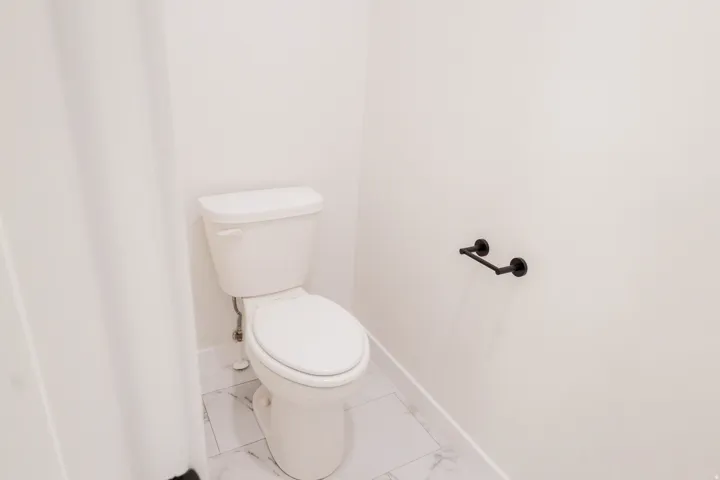 Bathroom featuring a white toilet, marble-look tile flooring, white baseboards, and a matte black wall-mounted fixture