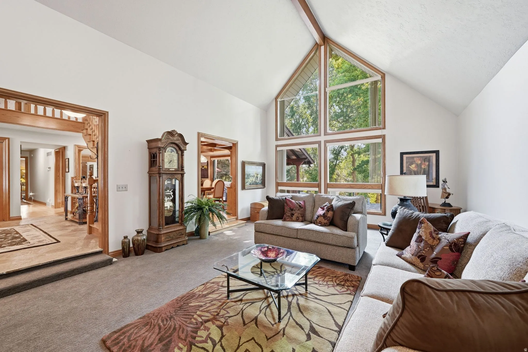 Vaulted ceiling living area featuring a prominent wall of windows with wood trim, a light-toned carpet, and a glass-top coffee table