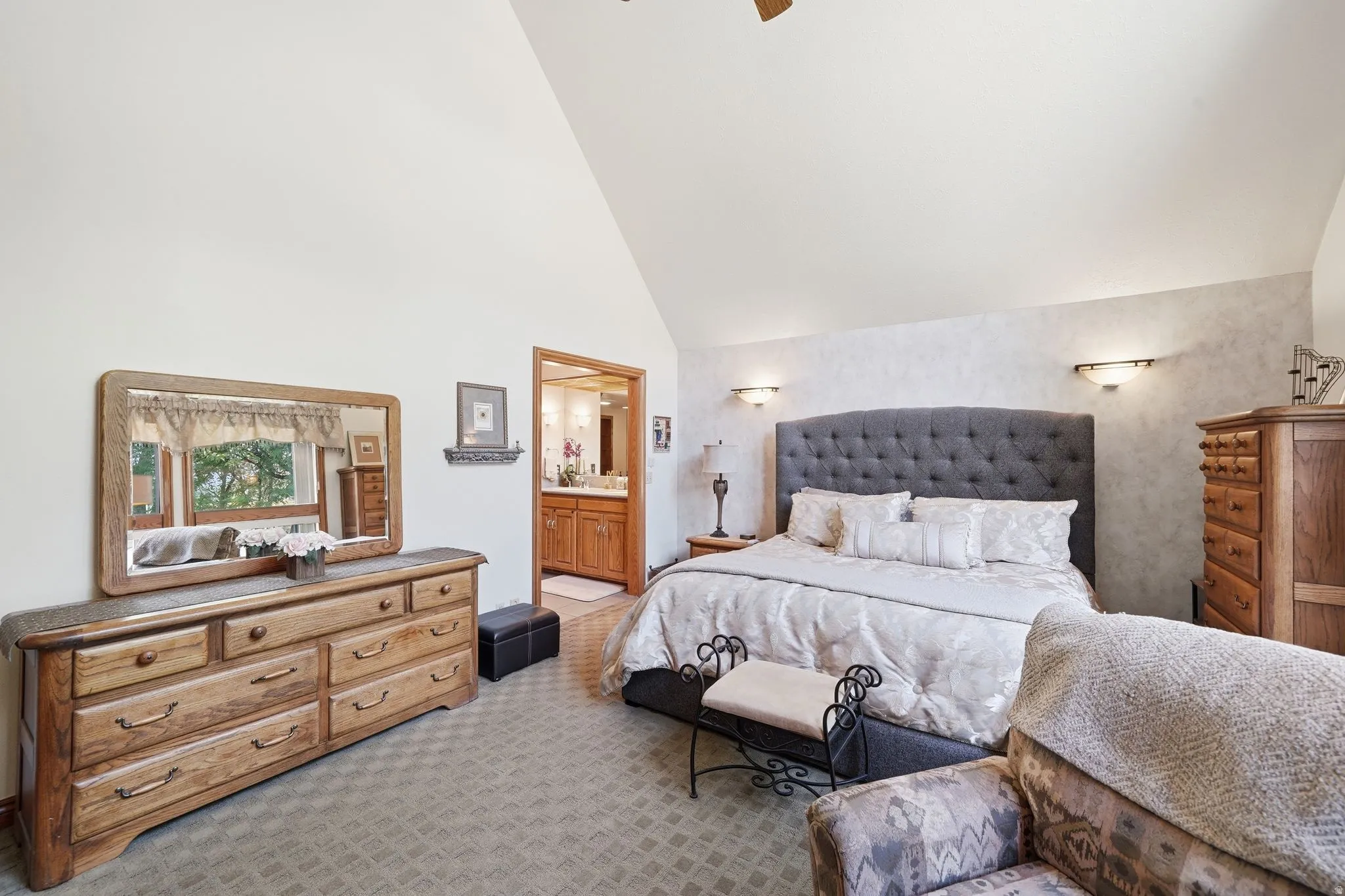 Vaulted ceiling bedroom with integrated wall sconces, textured wall treatment, and an open doorway revealing a bathroom with wood cabinetry and a double vanity