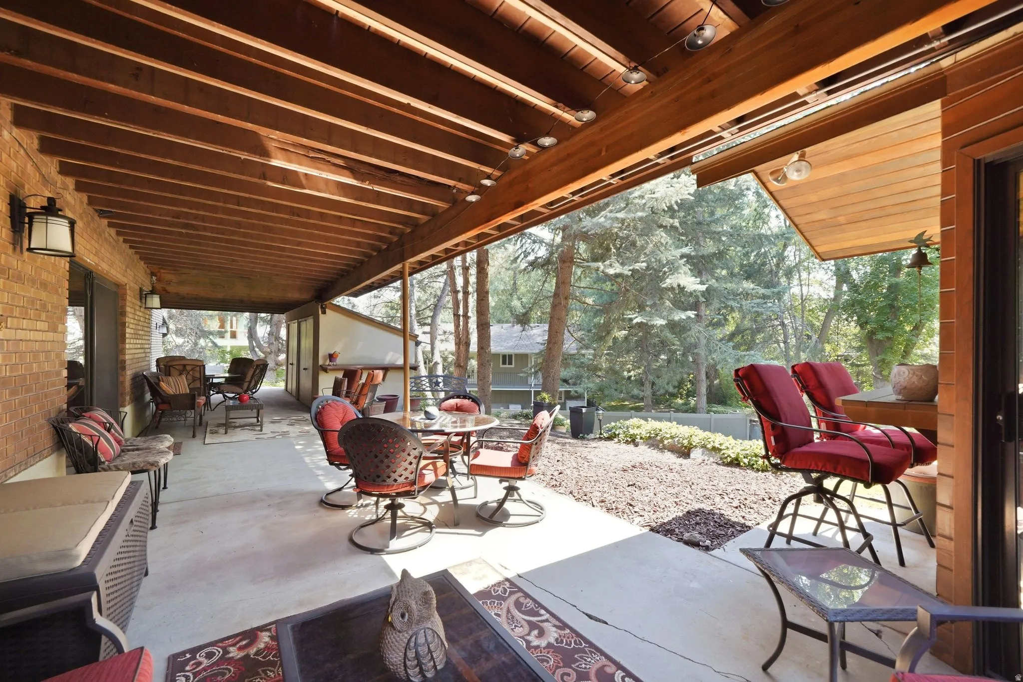 Expansive covered patio featuring a concrete surface, exposed wood beam ceiling, brick exterior wall, and outdoor lighting fixtures