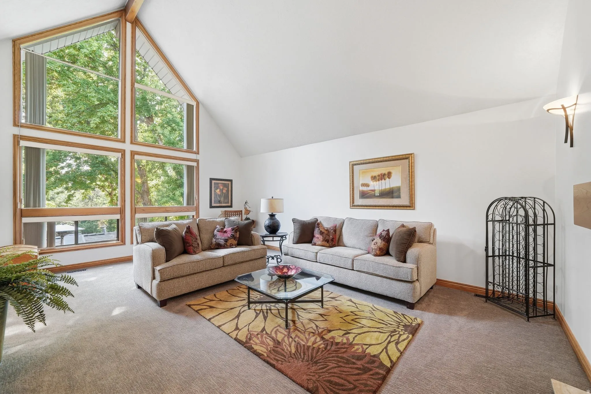 Vaulted ceiling living area featuring a prominent full-height window wall with wood trim, wall-mounted sconce lighting, and soft carpet flooring