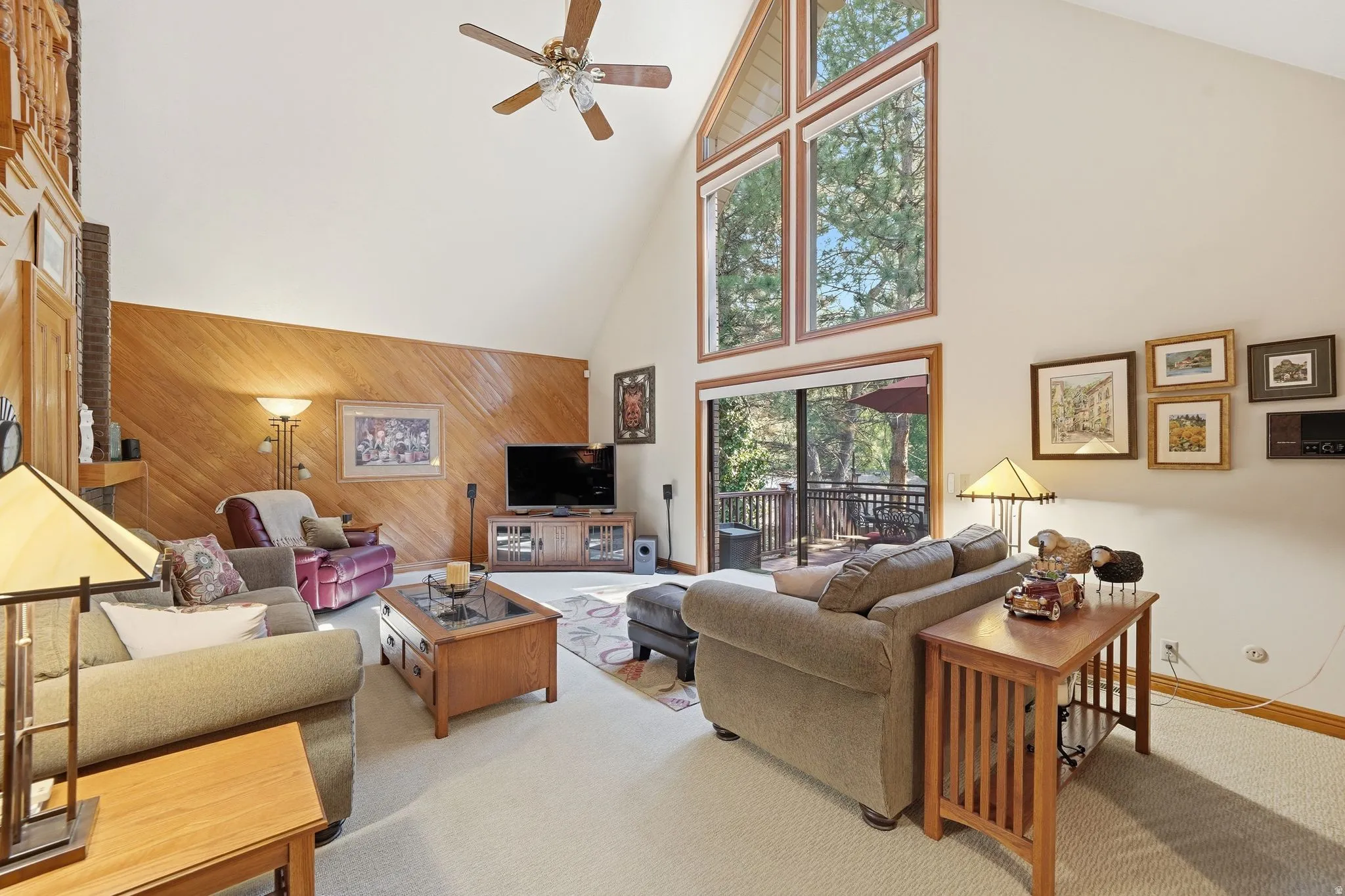 Vaulted ceiling living space featuring a ceiling fan, wood-paneled accent wall, a sliding glass door, and large windows