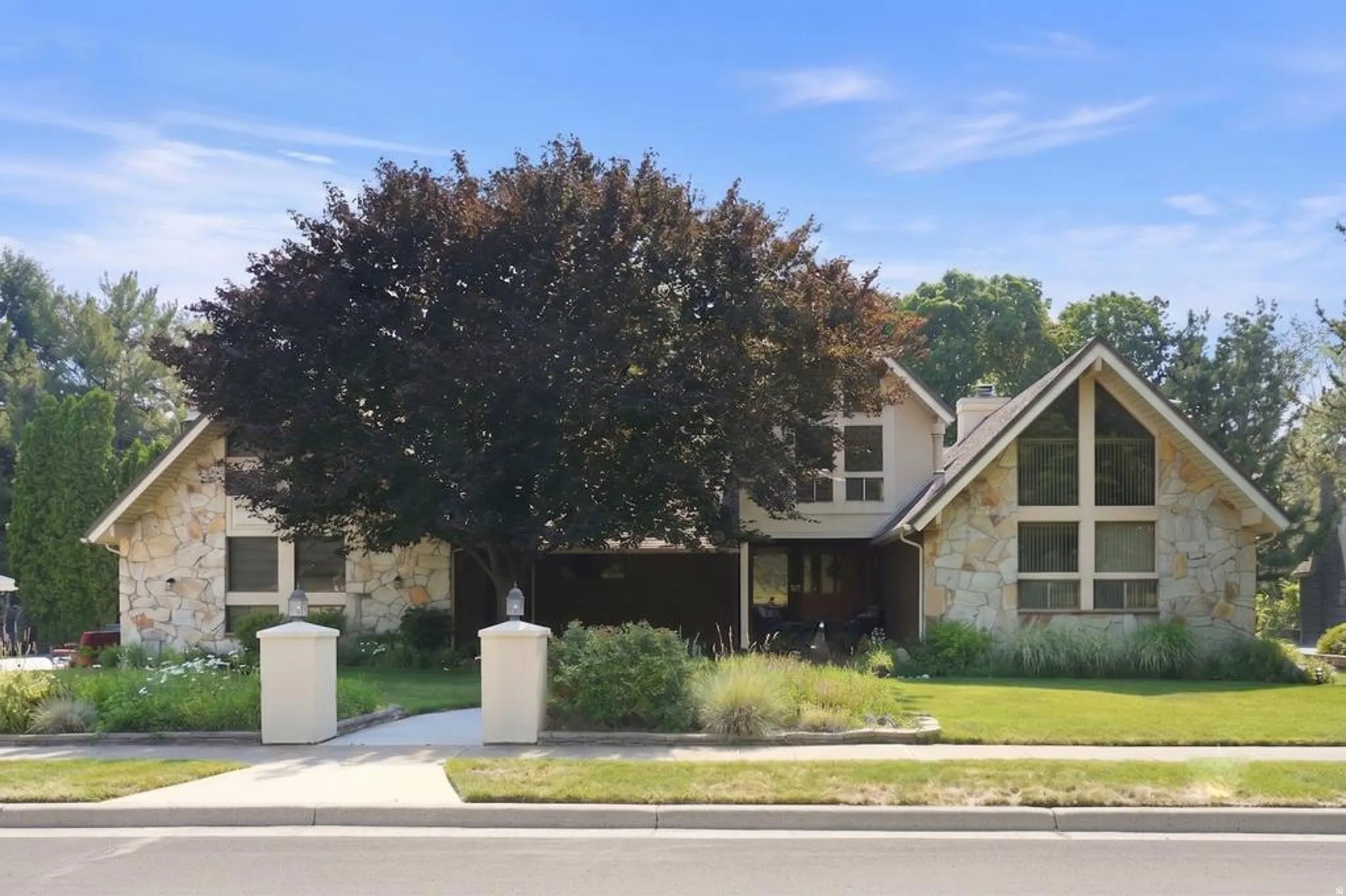 Stone facade residence featuring a prominent gabled roofline, expansive front windows, a mature front yard tree, and an inviting walkway flanked by decorative pillars