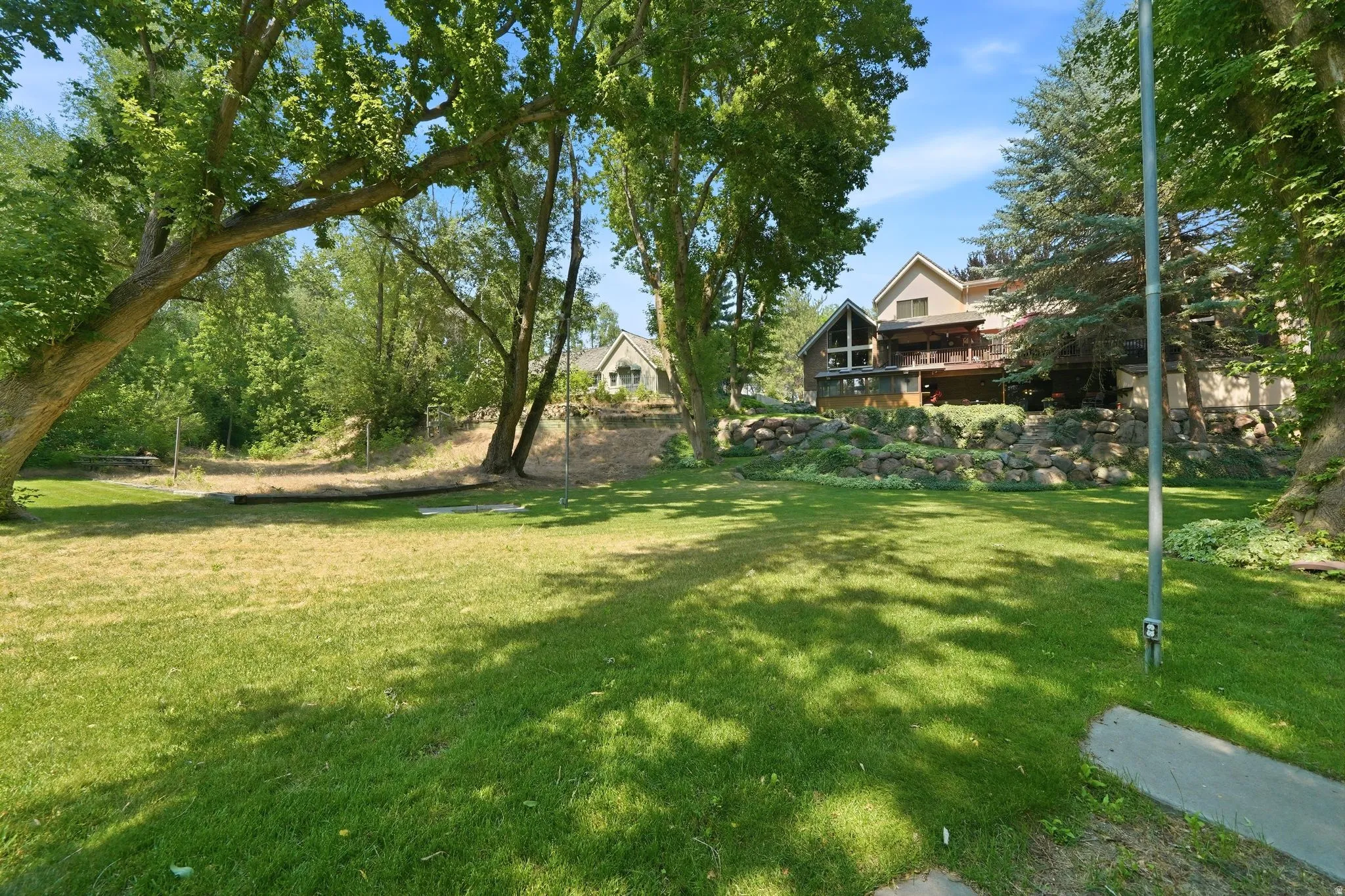 Expansive green lawn with mature trees and a two-story residence featuring a large deck and rock landscaping