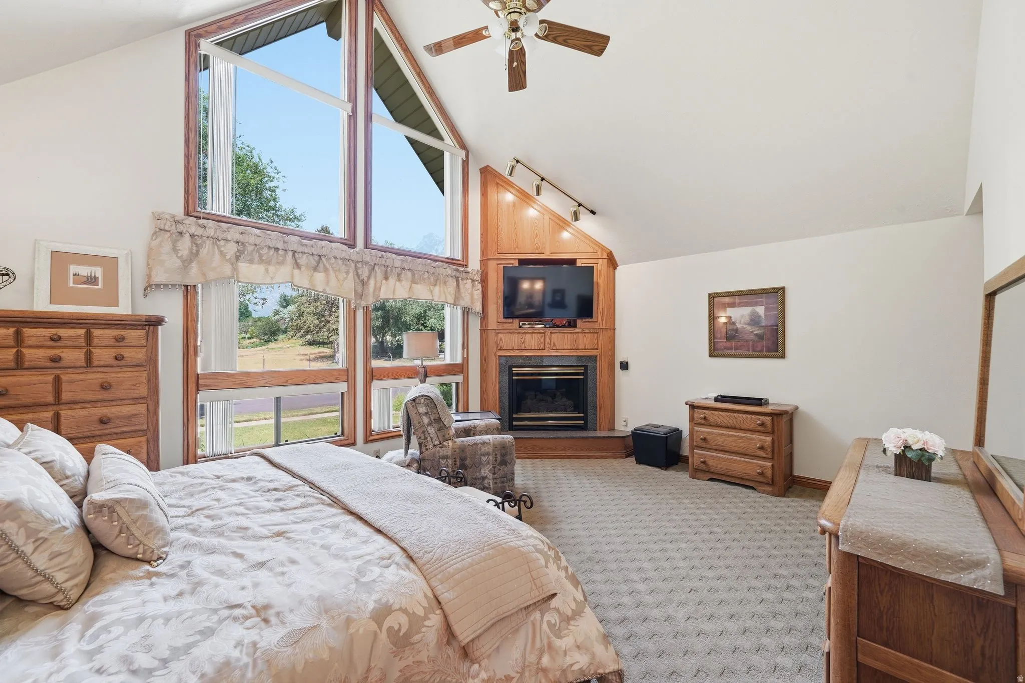 Vaulted ceiling bedroom featuring expansive windows, a wood-finish fireplace surround, and a ceiling fan