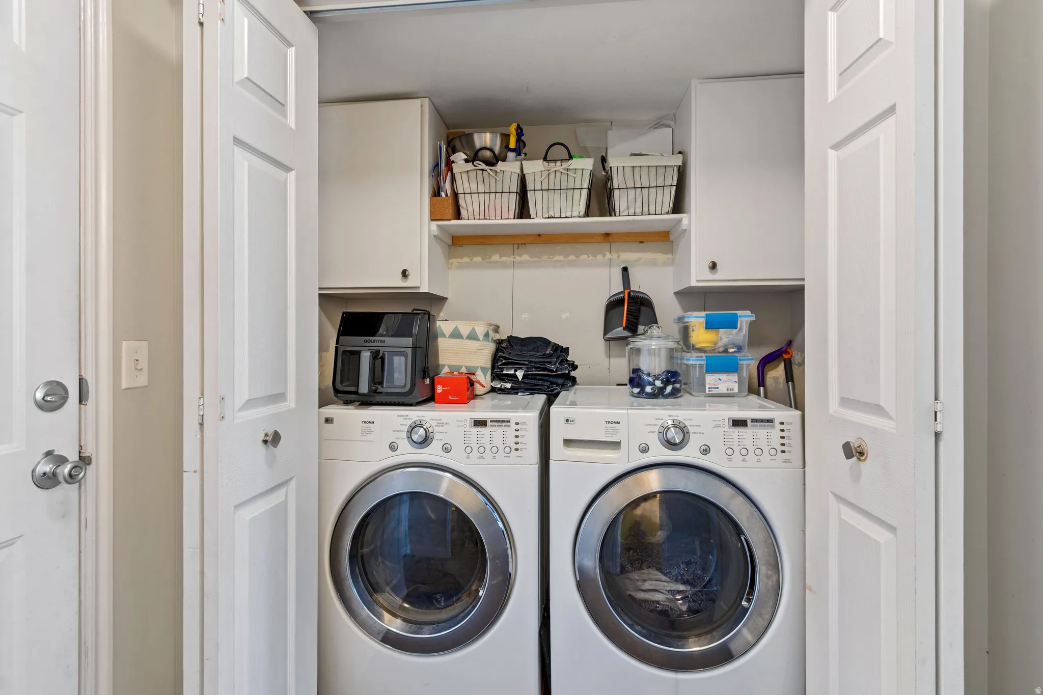 Dedicated laundry area featuring white bi-fold doors, upper cabinetry, an open shelf, and white front-load appliances