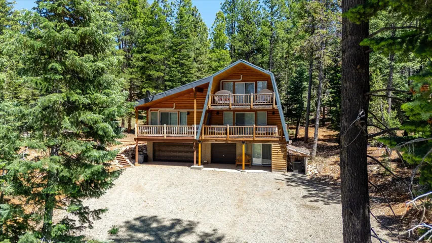 Cedar-sided A-frame residence featuring multiple balconies with white railings, a gravel driveway, and surrounding mature conifer trees