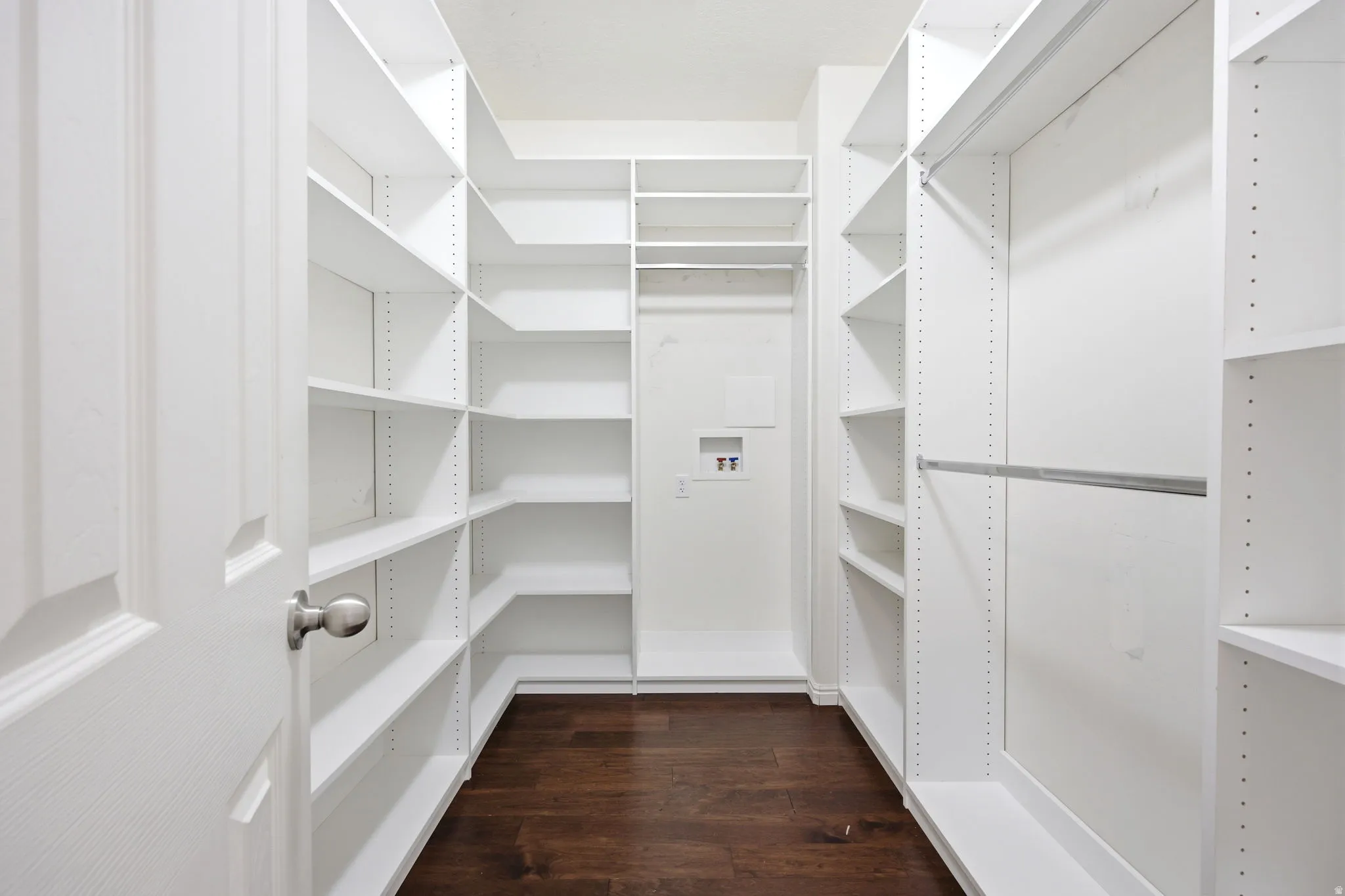 Spacious walk-in closet featuring extensive white built-in shelving, multiple hanging rods, dark wood-finish flooring, and integrated laundry hookups