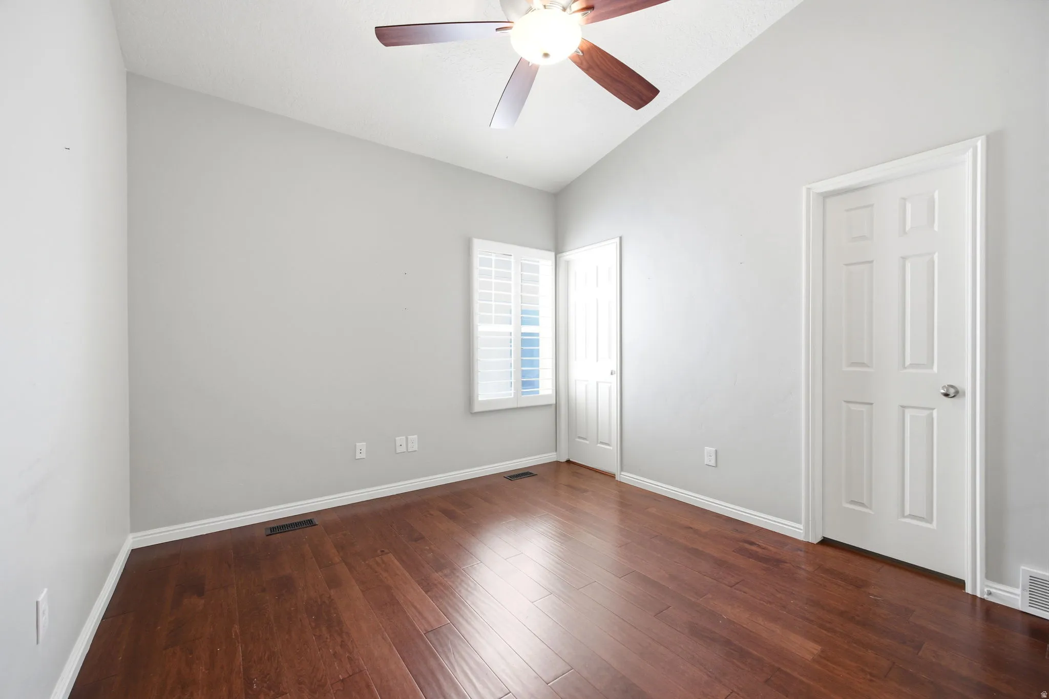 Sloped ceiling room featuring wood-finish flooring, a ceiling fan with light fixture, white plantation shutters, and white paneled doors