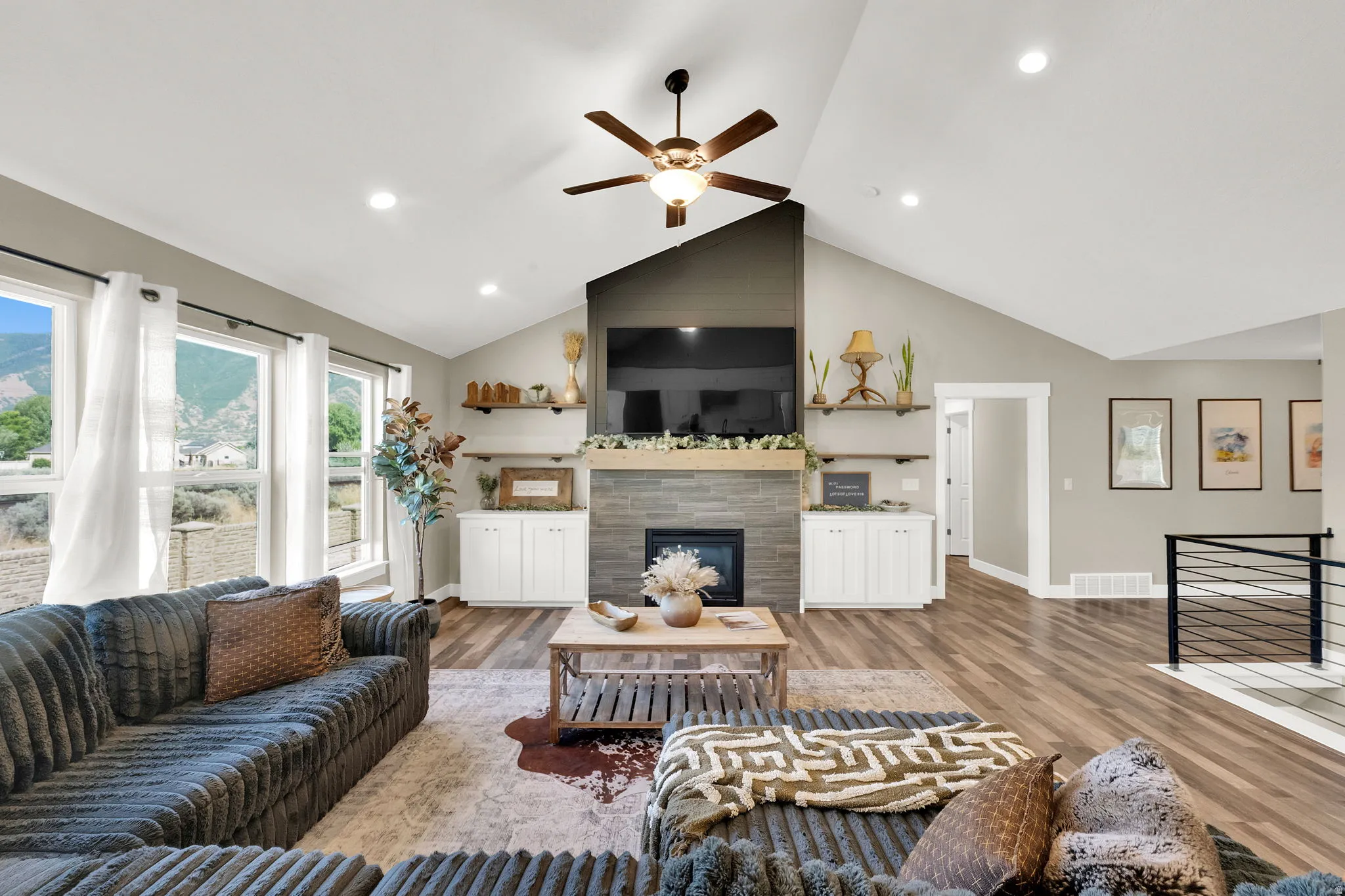 Spacious living area featuring a vaulted ceiling with a ceiling fan, a stone-clad fireplace with a wood mantel, built-in cabinetry, and wood-finish flooring