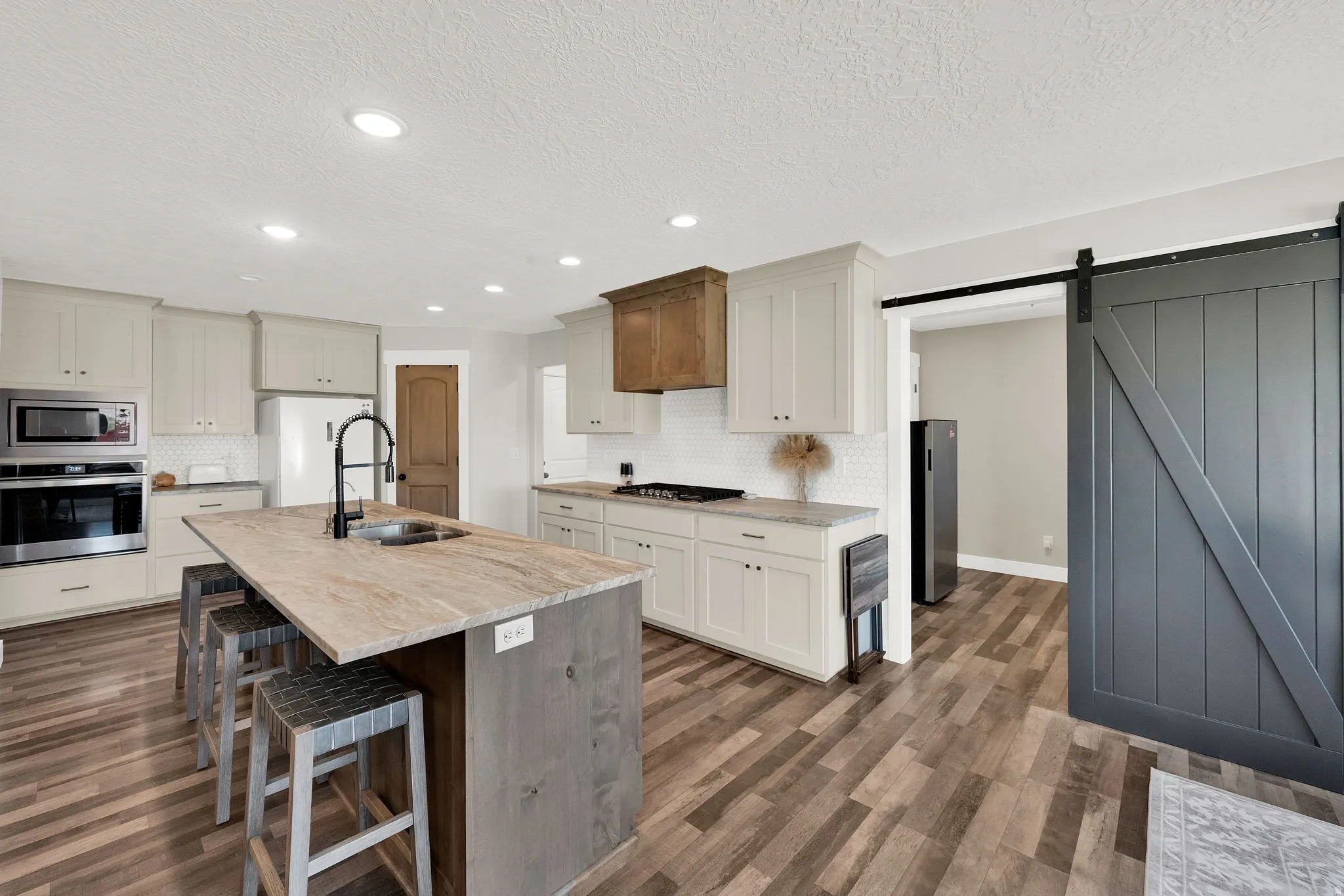 Kitchen featuring a large island with an integrated sink, wood-finish flooring, white subway tile backsplash, stainless steel appliances, and a dark barn door