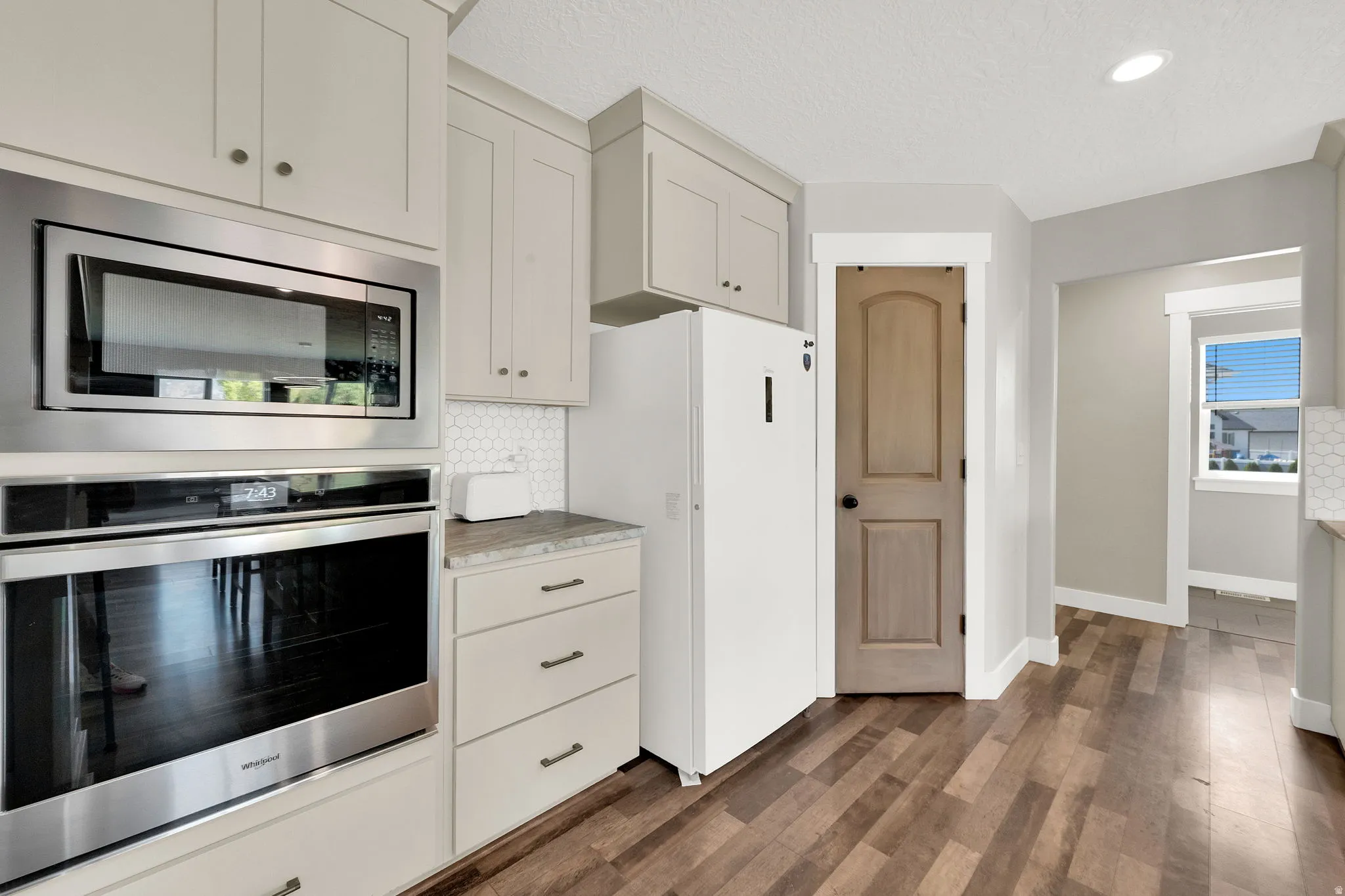 Kitchen featuring stainless steel built-in microwave and oven, light-toned cabinetry, hexagonal tile backsplash, wood-finish flooring, and a window providing natural light