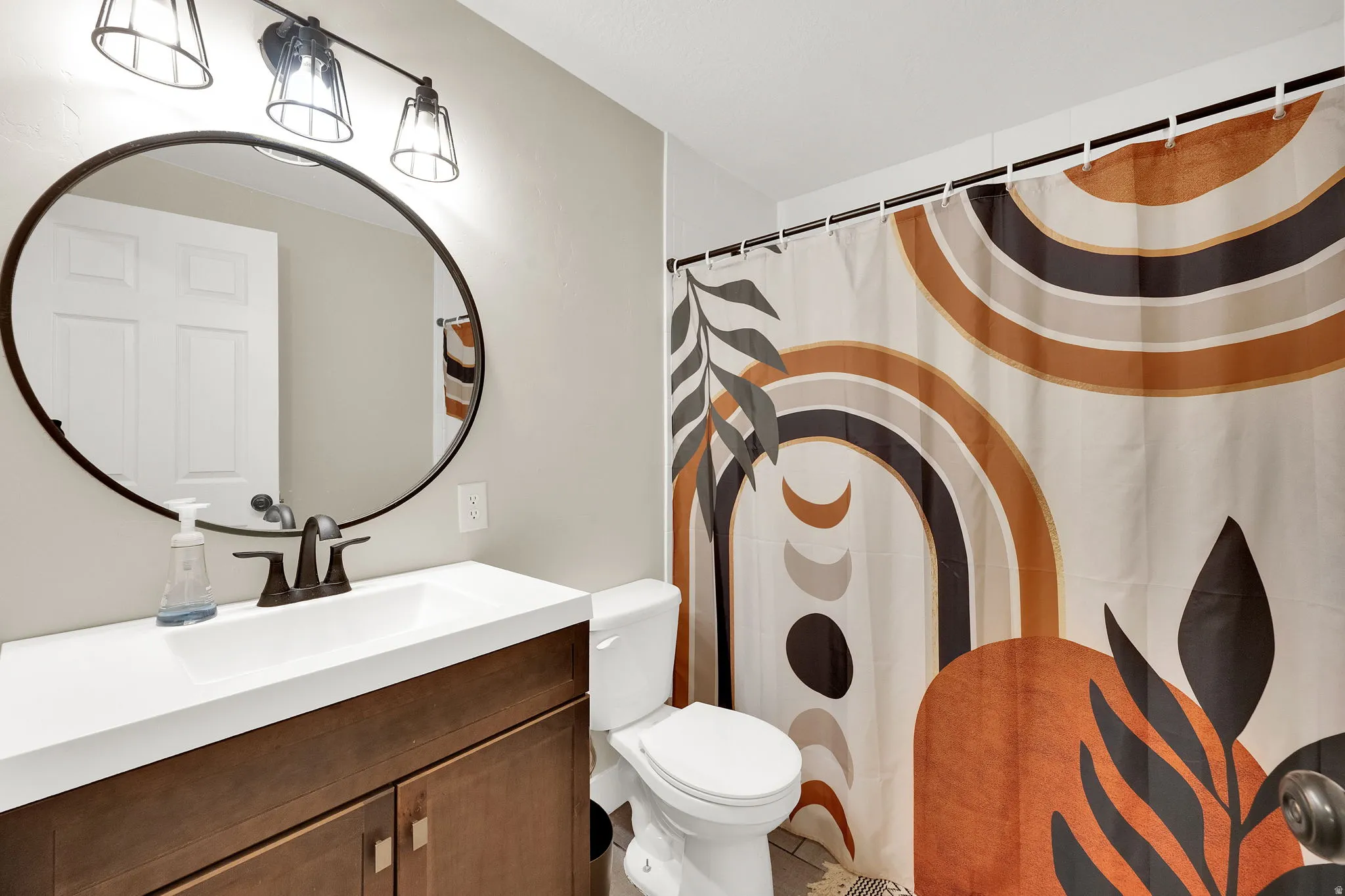 Bathroom featuring a wood-finish vanity with a white countertop and integrated sink, a dark-framed round mirror, and a three-light fixture with dark metal accents