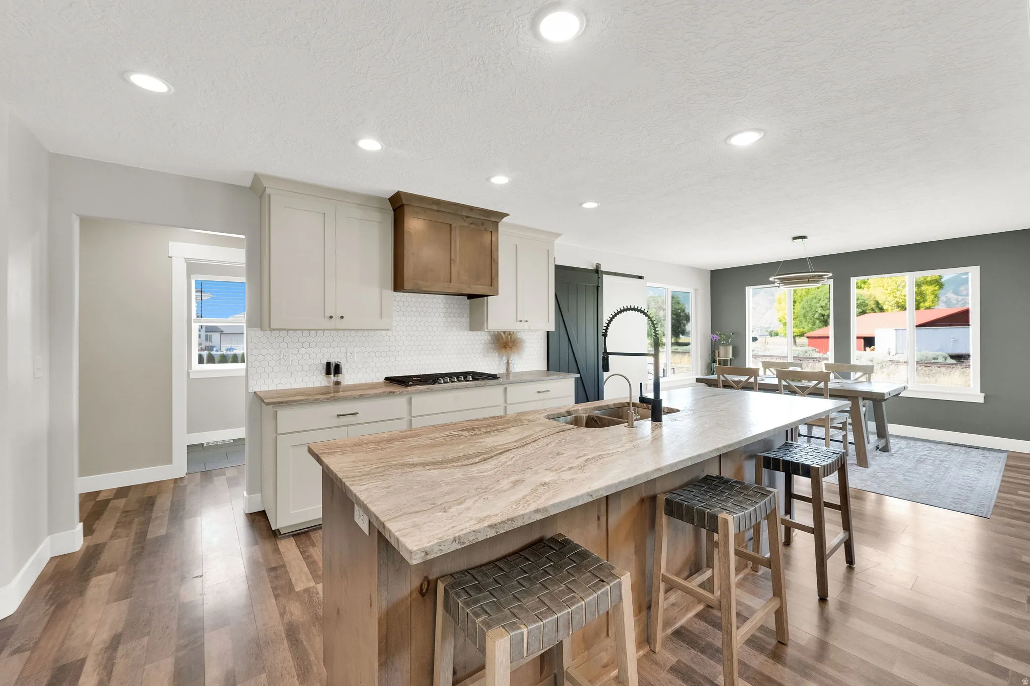 Spacious kitchen featuring a large island with stone countertop, wood-finish flooring, white cabinetry, a dark wood range hood, and a white hexagonal tile backsplash
