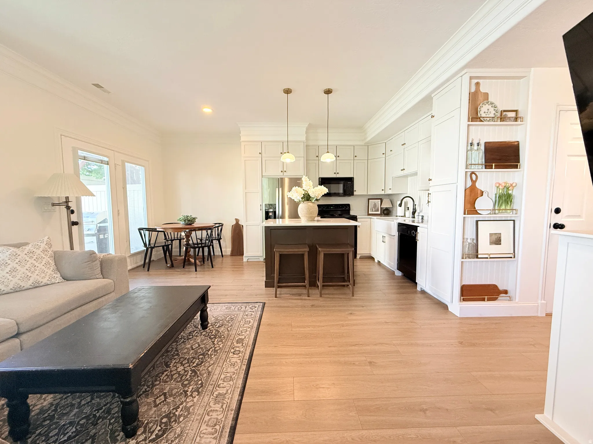 Open-concept living area featuring light wood-finish flooring, white upper cabinetry, a dark island with seating, a kitchen sink, and a built-in shelving unit
