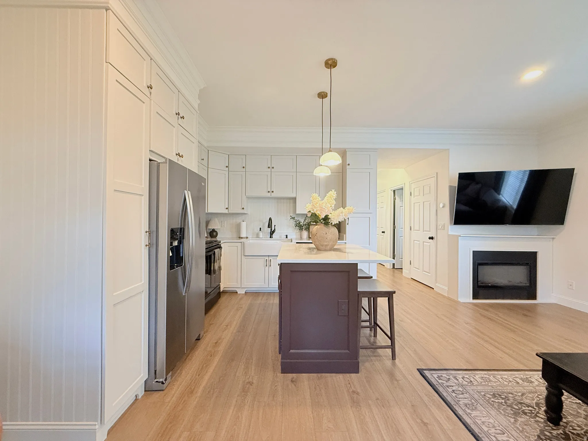 Open-concept living area featuring light wood-finish flooring, a white kitchen with a farmhouse sink, stainless steel appliances, and a central island with counter seating