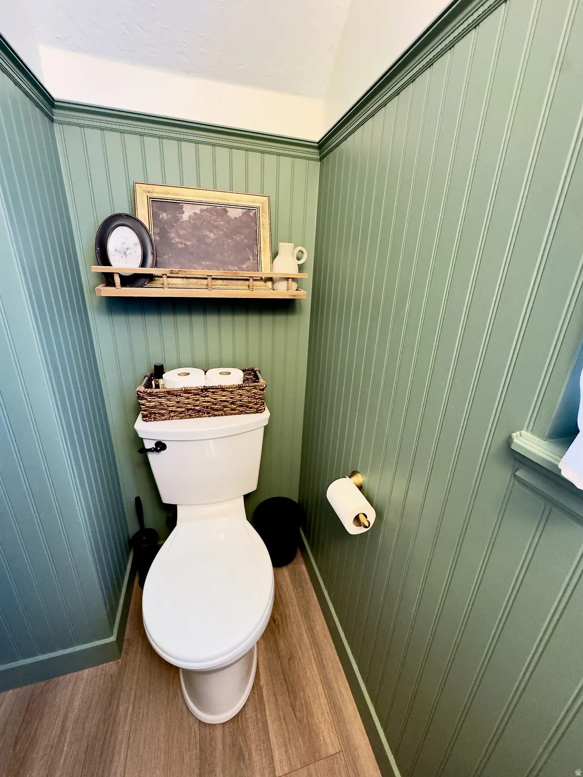 Bathroom featuring a white toilet, gold-toned toilet paper holder, wood-finish flooring, and green beadboard walls with white trim