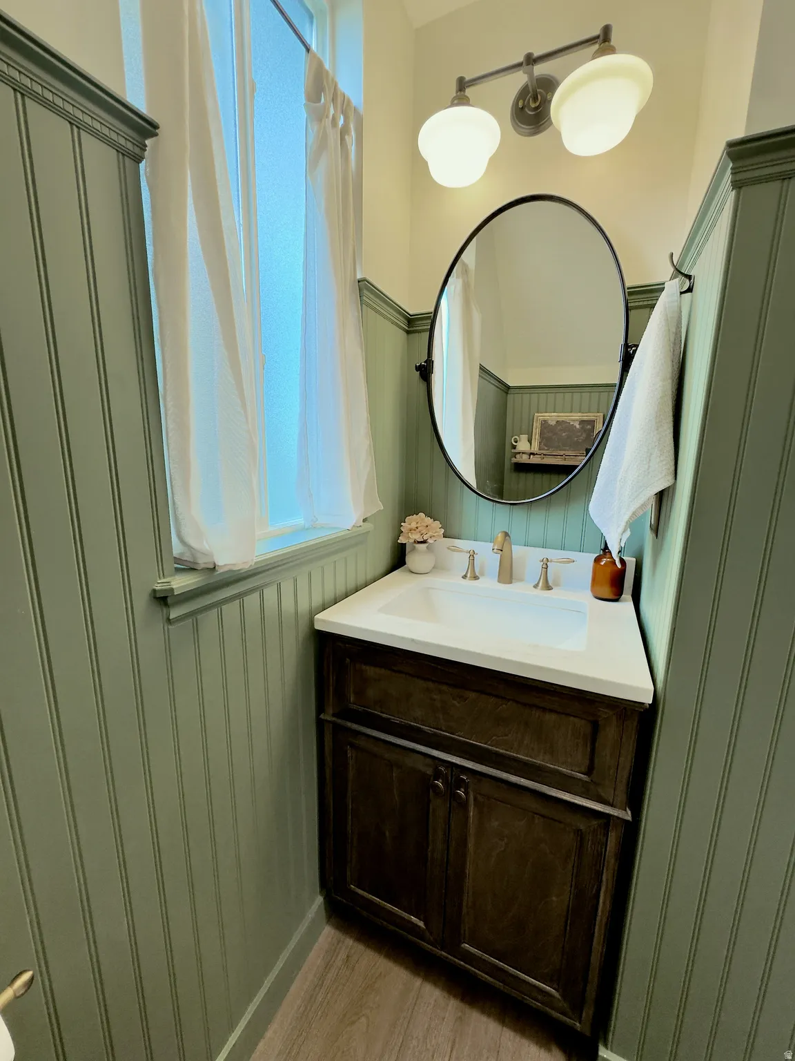 Bathroom featuring beadboard wainscoting, a dark wood-finish vanity with a white countertop, and a contemporary oval mirror
