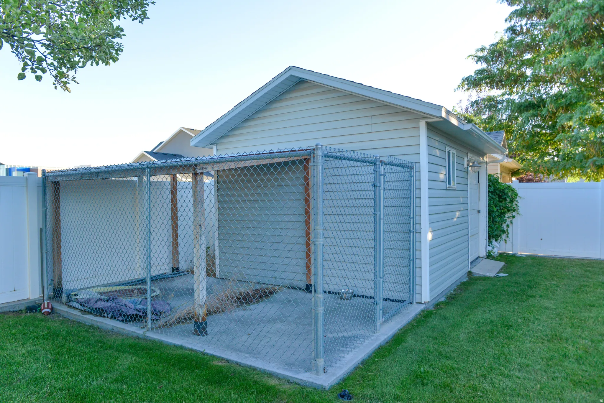 Detached shed featuring light gray siding, white trim, and a shingled roof, accompanied by an attached chain-link enclosure with a concrete pad