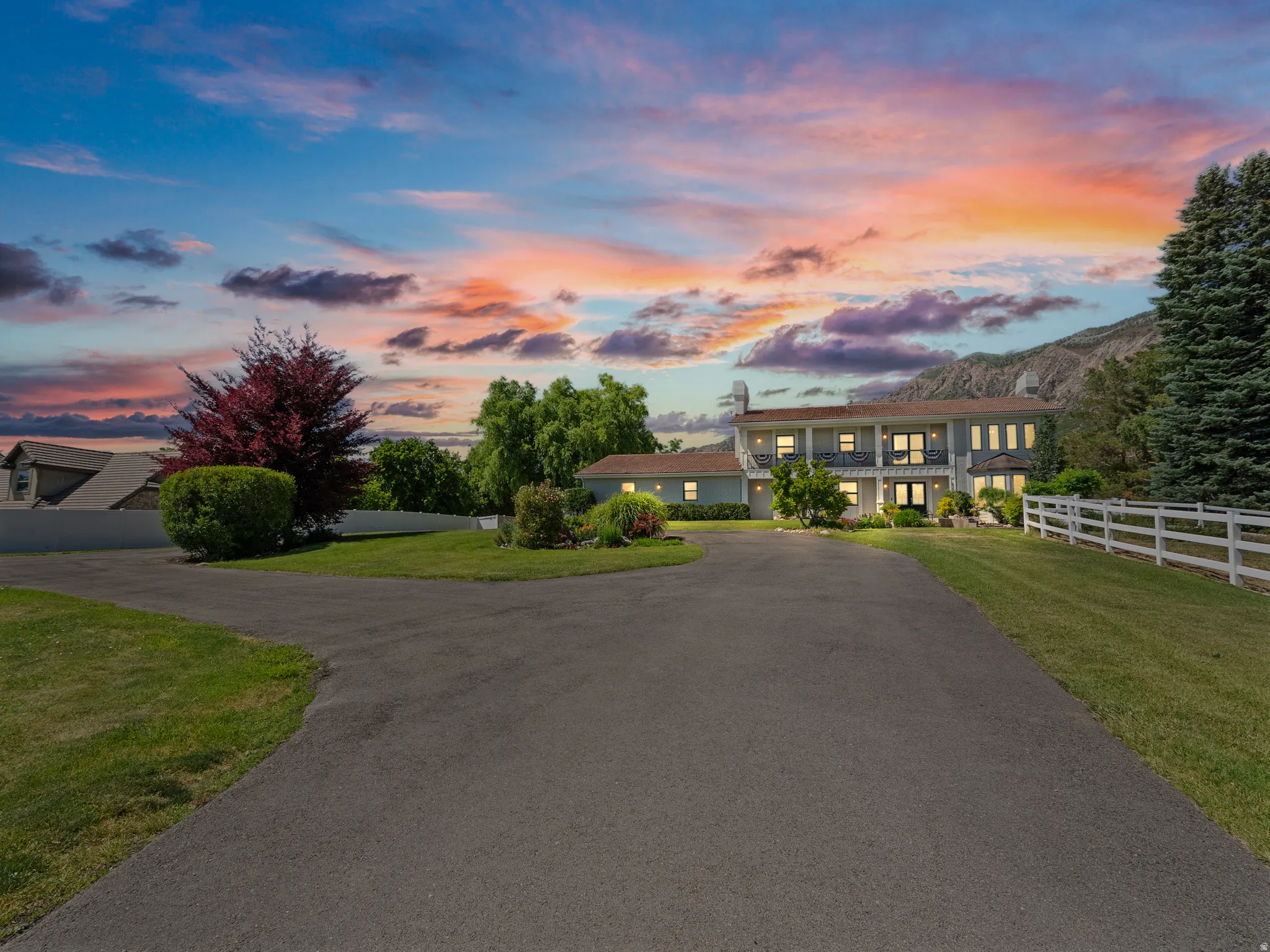 Asphalt driveway leading to a two-story residence with a multi-pane front door, exterior lighting, upper-level balcony, and a white picket fence