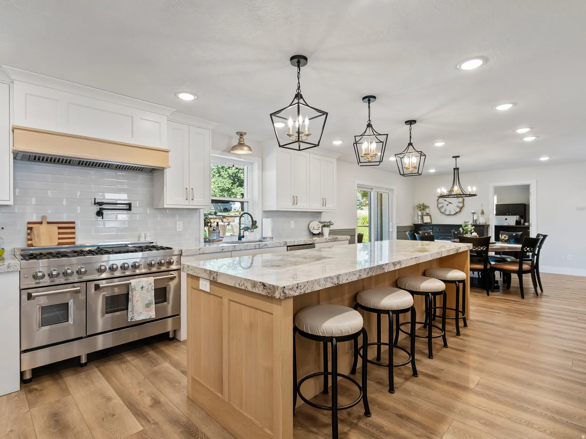 Open-concept kitchen featuring an expansive island with seating, stainless steel professional-grade range, light-toned wood-finish flooring, white shaker cabinetry, and recessed lighting