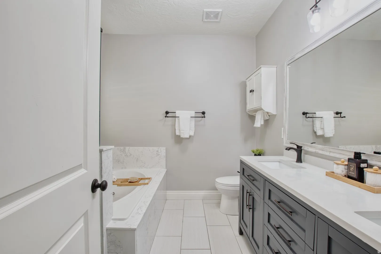Contemporary bathroom featuring a built-in soaking tub with a tiled surround, a dual vanity with a white countertop and dark cabinetry, a wall-mounted medicine cabinet, and white tile flooring