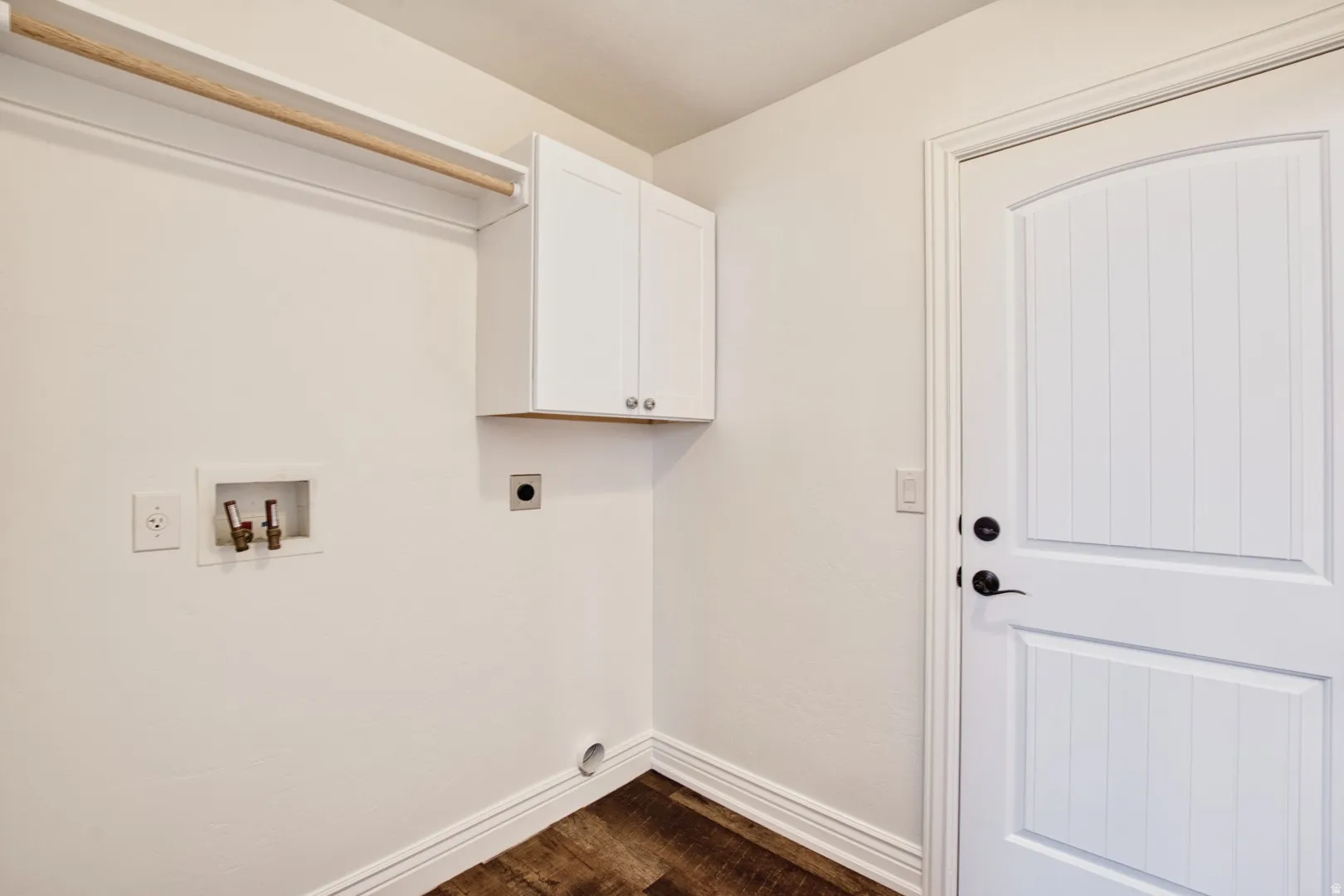 Dedicated laundry space featuring wood-finish flooring, white wall-mounted cabinetry, a built-in hanging rod, and a beadboard-style interior door with matte black hardware