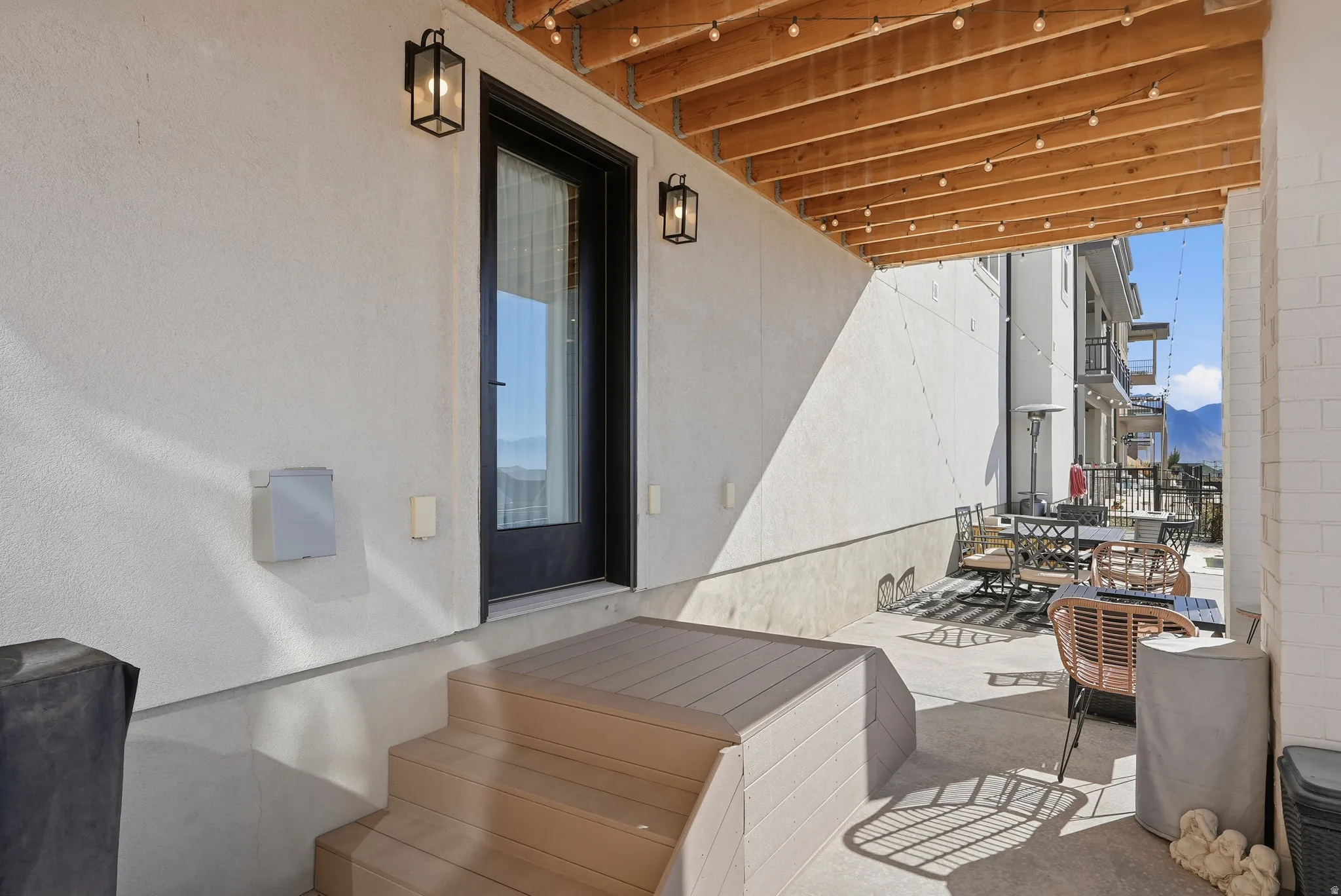 Covered patio featuring a wood-finish ceiling with string lights, a full-lite exterior door, two exterior wall lanterns, composite decking stairs, and a concrete patio surface