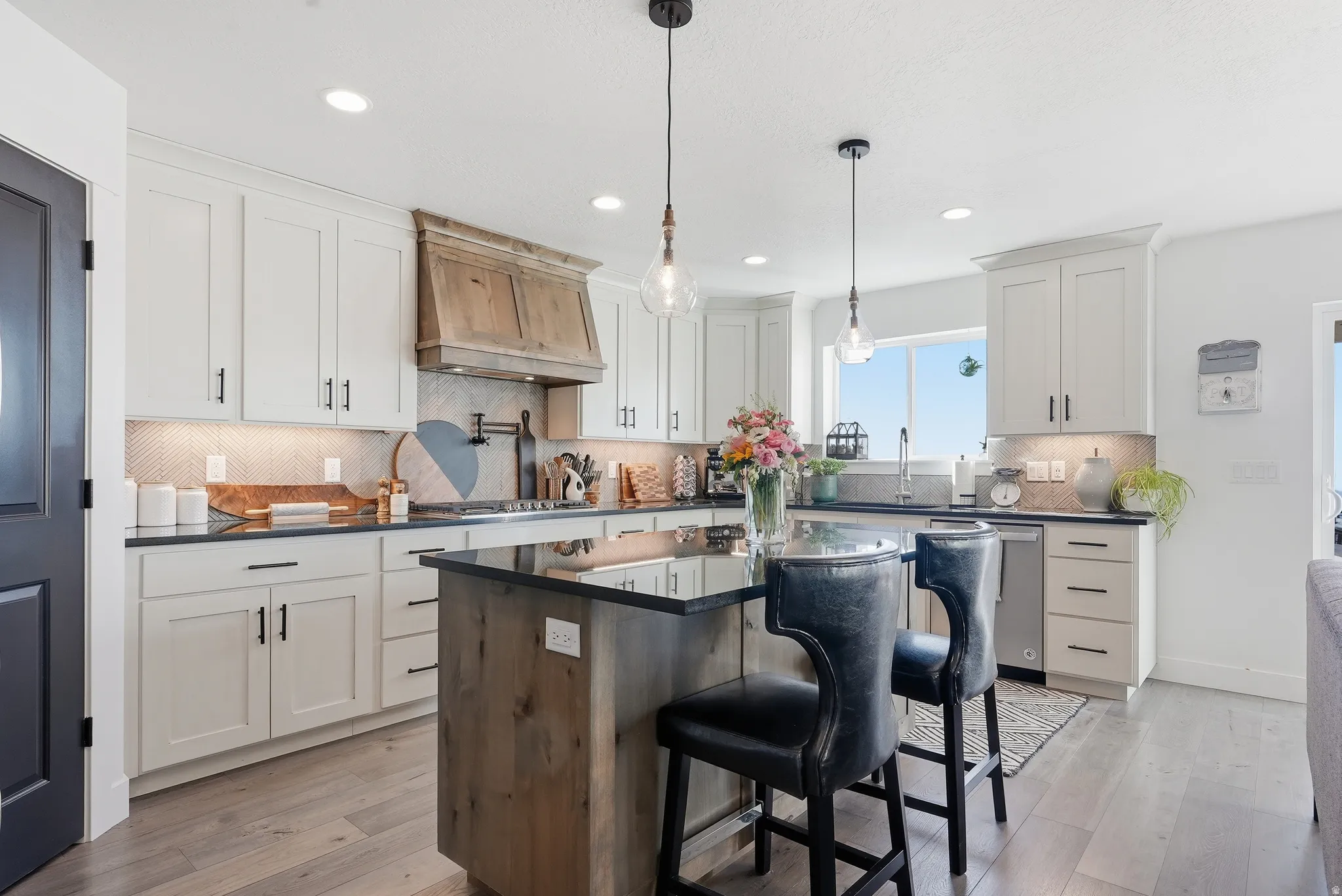 Kitchen featuring a dark countertop island with pendant lighting, white cabinetry, a wood-finish range hood, tile backsplash, and light wood-finish flooring