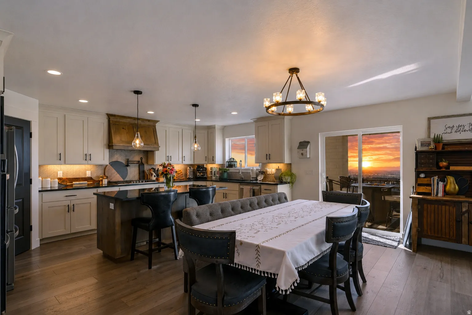 Open-concept kitchen and dining area featuring light-toned cabinetry, dark countertops, wood-finish flooring, a central island with seating, and sliding glass doors opening to a scenic sunset