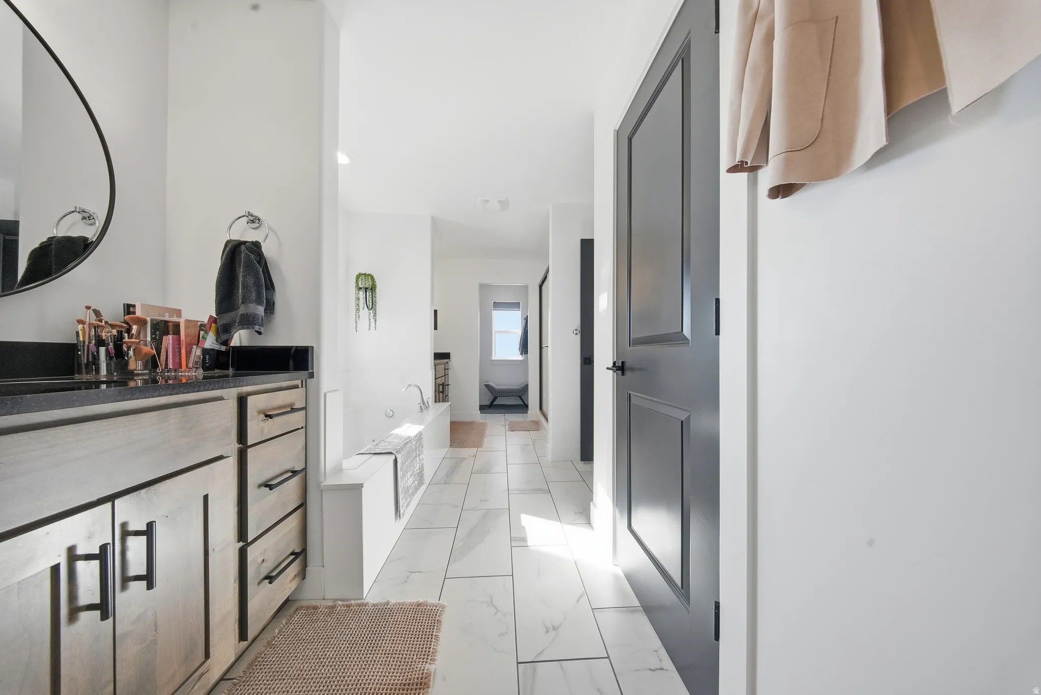Spacious bathroom featuring a light wood-finish vanity with dark countertop, large circular mirror, white tiled flooring, and a built-in soaking tub