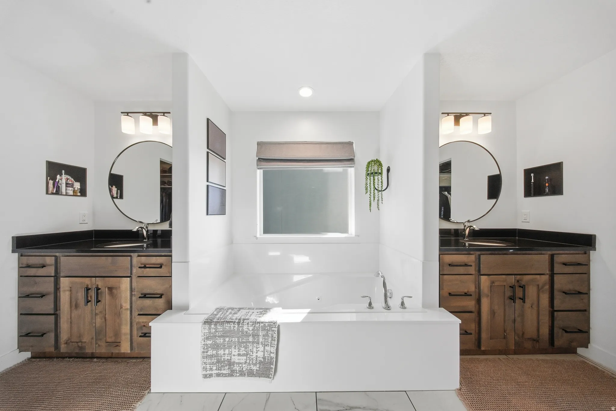 Soaking tub with surrounding white tile, framed by two wood-finish vanities with dark countertops and circular mirrors