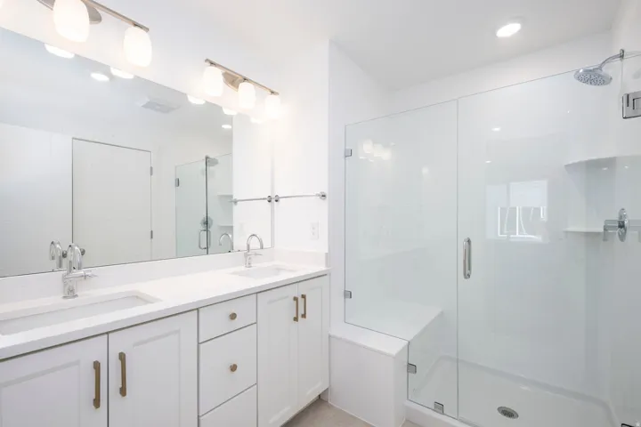 Contemporary bathroom featuring a spacious glass-enclosed shower with a built-in bench, dual vanity with undermount sinks, and white cabinetry with brass hardware