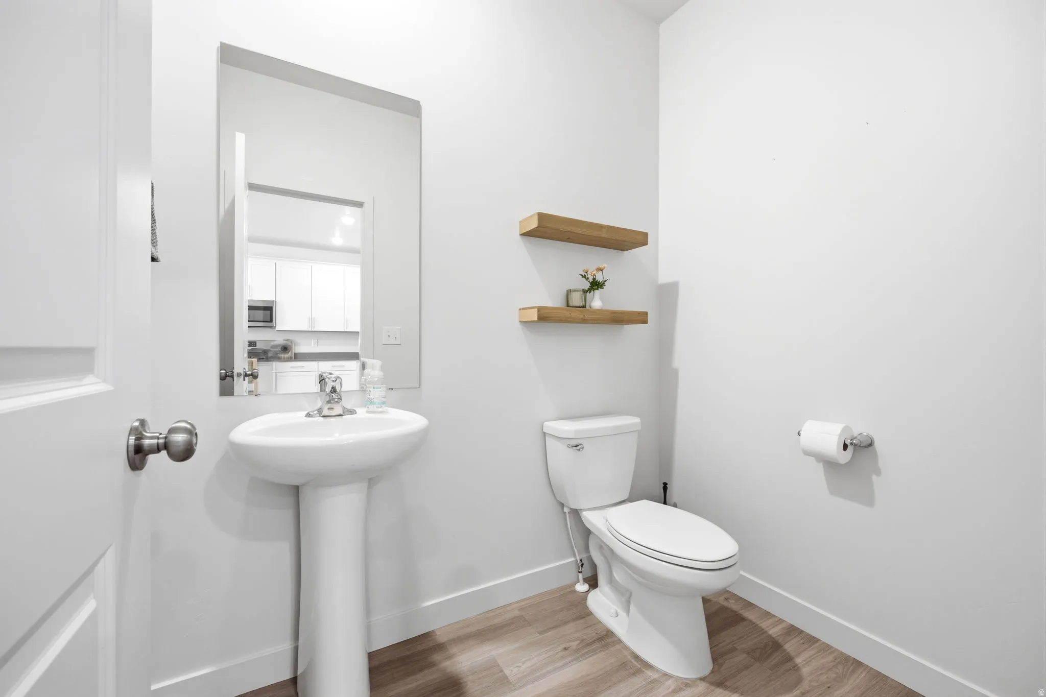 Compact powder room featuring a pedestal sink, a wall-mounted mirror, two floating wood-finish shelves, and wood-finish flooring