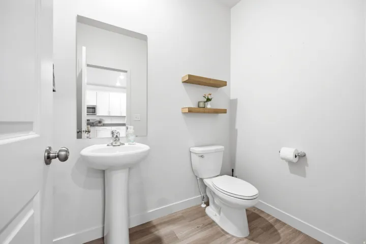 Compact powder room featuring a pedestal sink, a wall-mounted mirror, two floating wood-finish shelves, and wood-finish flooring