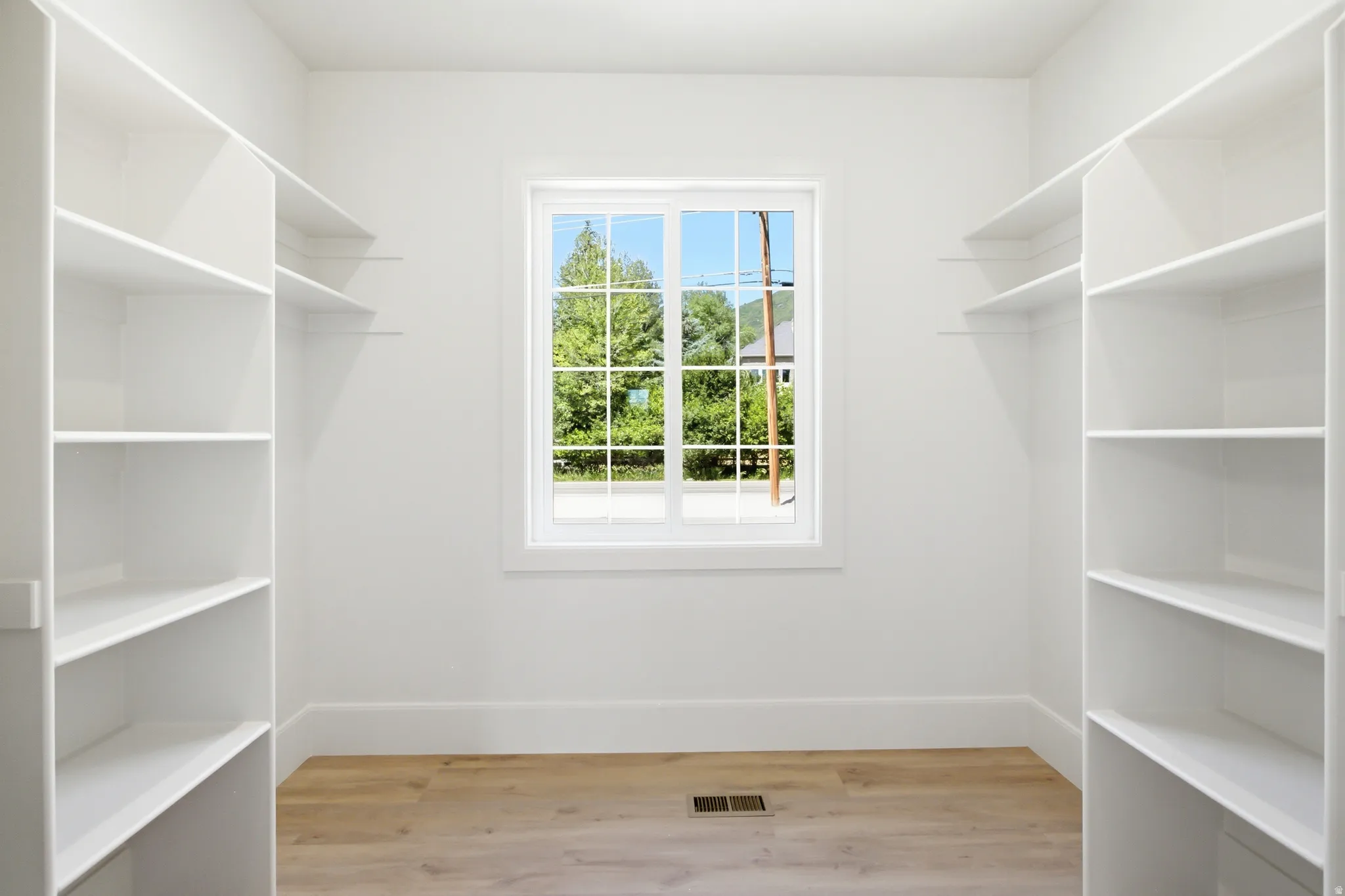Walk-in closet featuring white built-in shelving, light wood-finish flooring, and a central window providing natural illumination
